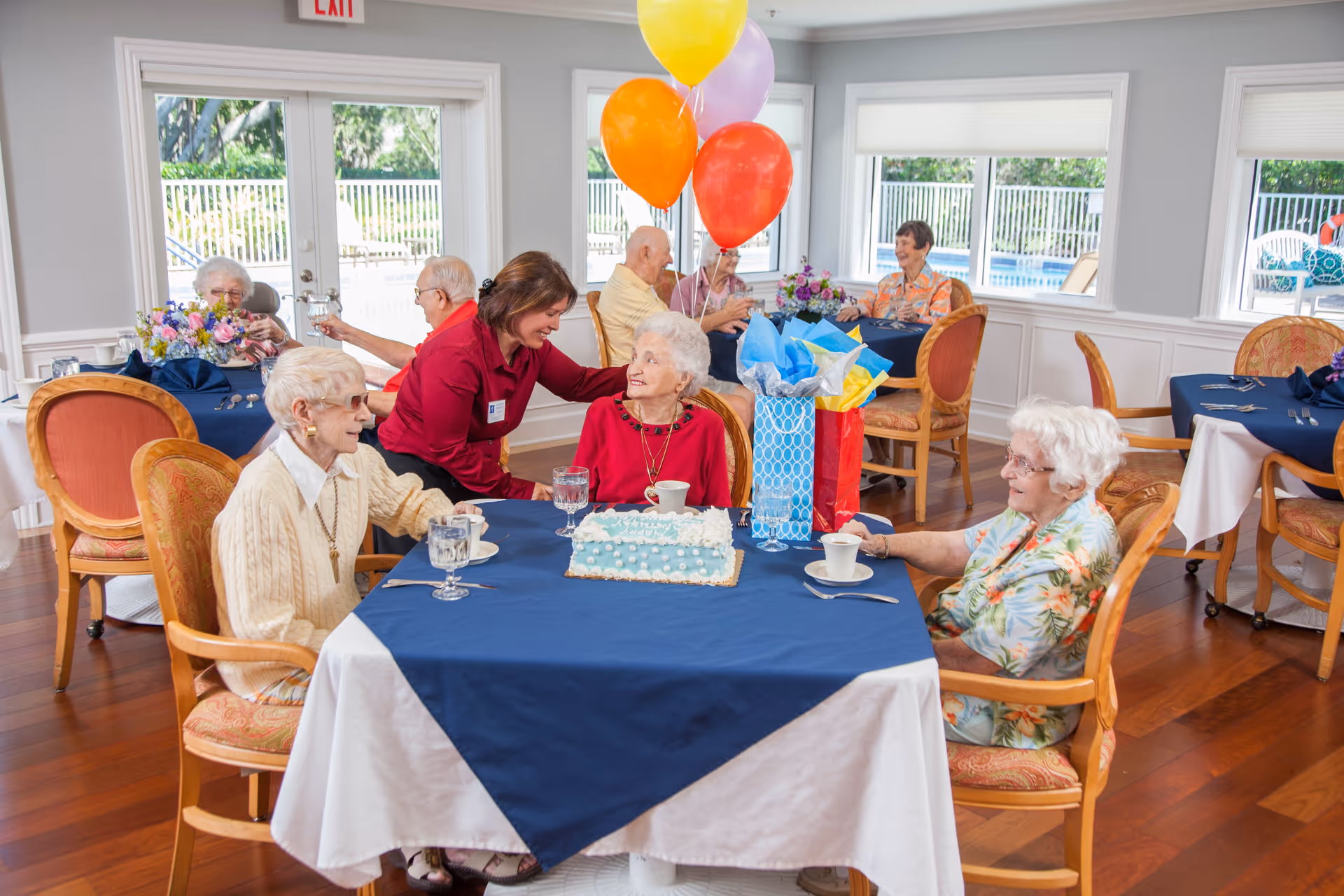 A group of elderly women sitting around a table in a dining room decorated with balloons and a birthday cake. A staff member in a red shirt is interacting warmly with one of the women. The room has large windows showing an outdoor pool area and is furnished with wooden chairs and tables covered with blue and white tablecloths.