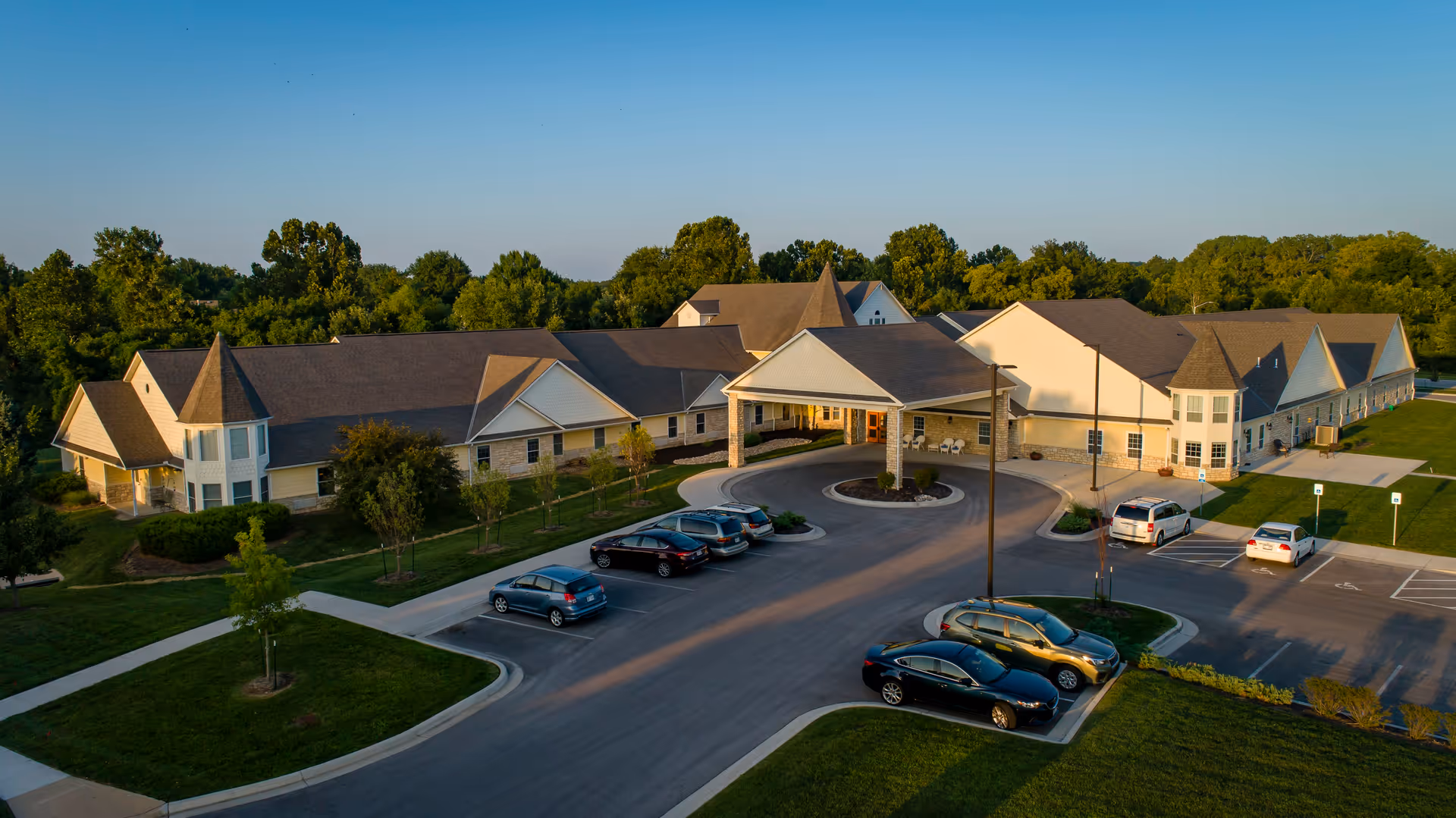 Aerial view of Homestead Assisted Living & Memory Care of Overland Park, showing a large single-story building with multiple peaked roofs and a covered entrance. Several cars are parked in the parking lot in front of the building, surrounded by green lawns and trees under a clear blue sky.