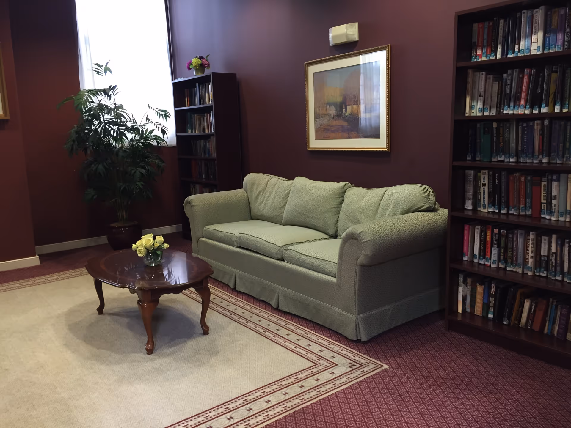 A cozy sitting area with a green upholstered sofa, a wooden coffee table with a small vase of yellow flowers, two tall bookshelves filled with books, a potted plant near a window with sheer curtains, and a framed painting on a maroon wall.