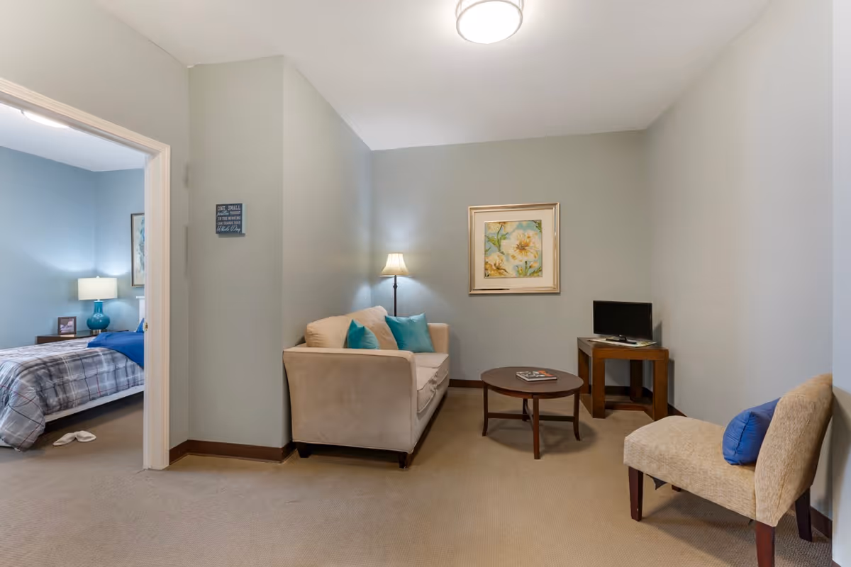 A cozy living room area with a beige sofa adorned with teal pillows, a round wooden coffee table with magazines, a floor lamp, a framed floral painting on the wall, and a small wooden side table holding a flat-screen TV. To the left, an open doorway reveals a bedroom with a bed covered in a plaid comforter, a nightstand with a blue lamp, and slippers on the floor.