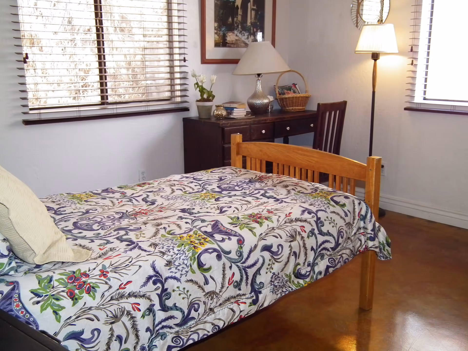 A bedroom with a wooden bed frame covered with a colorful floral patterned bedspread. There are two windows with blinds, a dark wooden desk with a lamp, books, a basket, and a chair. A floor lamp is also visible near the window.