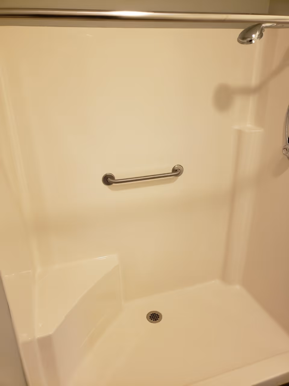 A clean walk-in shower with a built-in seat, stainless steel grab bar, shower rod, and handheld shower head.