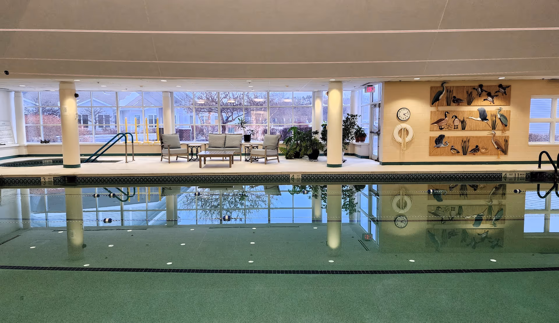 Indoor swimming pool area with clear water, surrounded by white columns and large windows letting in natural light. There are several cushioned chairs and a table on the pool deck, along with potted plants. On the far wall, there is a clock, a life preserver, and wooden wall art featuring various birds.