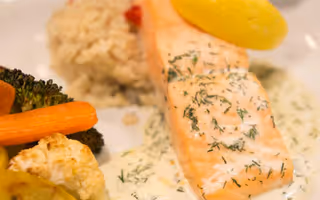 Close-up of a plated meal featuring a piece of cooked salmon topped with a creamy dill sauce and a lemon wedge, served with a side of rice and steamed mixed vegetables including carrots, broccoli, and cauliflower.