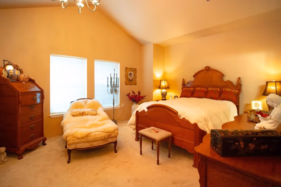 A warmly lit bedroom with a wooden bed featuring an ornate headboard and footboard, a small upholstered bench at the foot of the bed, a chaise lounge covered with a fluffy white throw, two windows with blinds, a wooden dresser with decorative items, a candelabra, and two table lamps providing soft lighting.