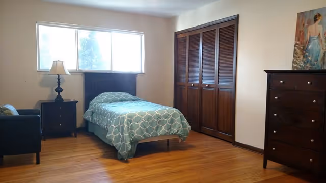 A simple bedroom with a single bed covered in a blue patterned bedspread, a wooden nightstand with a lamp, a dark armchair, a wooden dresser, and a large closet with wooden louvered doors. There is a window letting in natural light and a painting on the wall above the dresser.