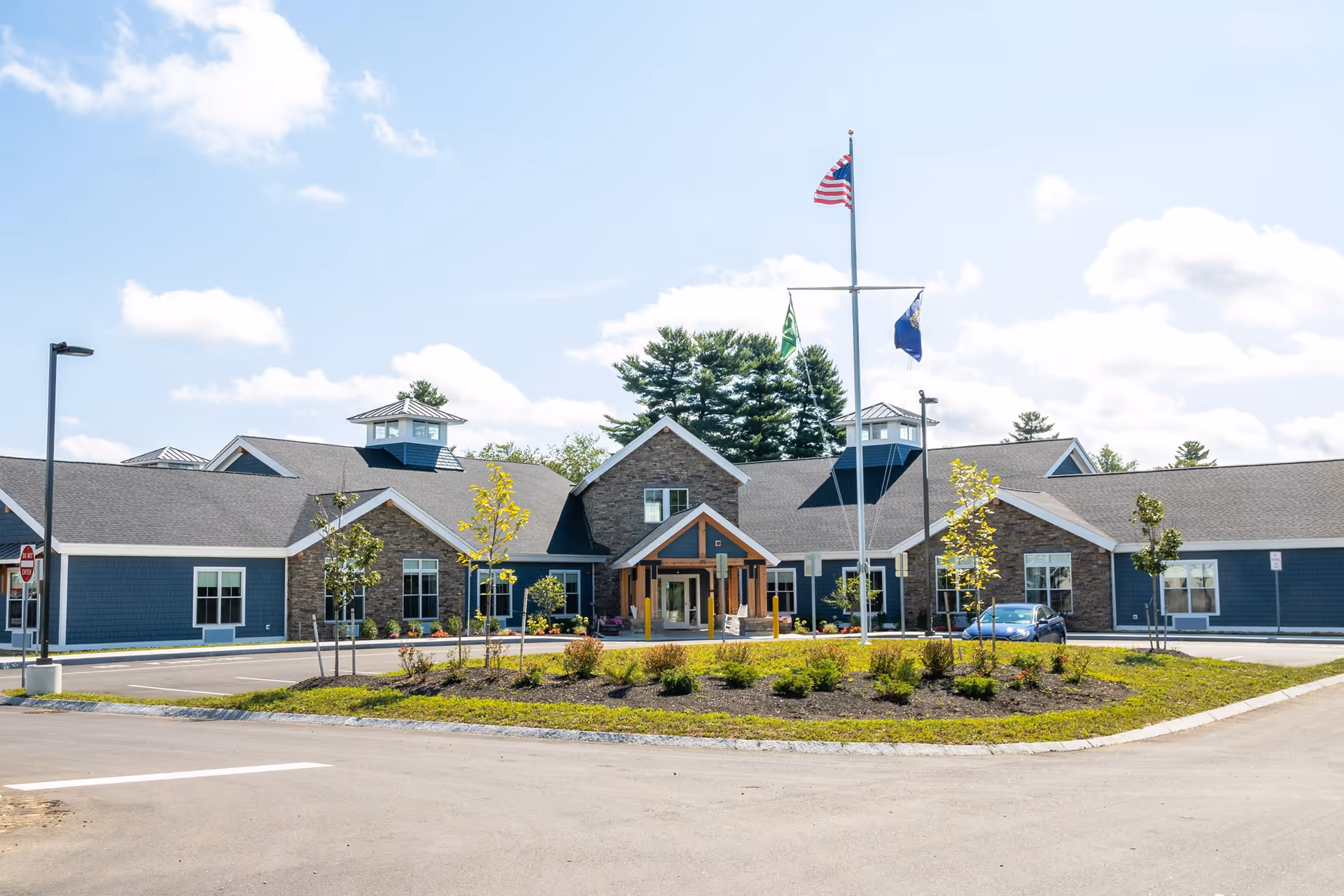 Front exterior view of a single-story senior living facility building with blue siding and stone accents, a covered entrance, landscaped greenery, and three flagpoles with flags including the American flag under a partly cloudy sky.