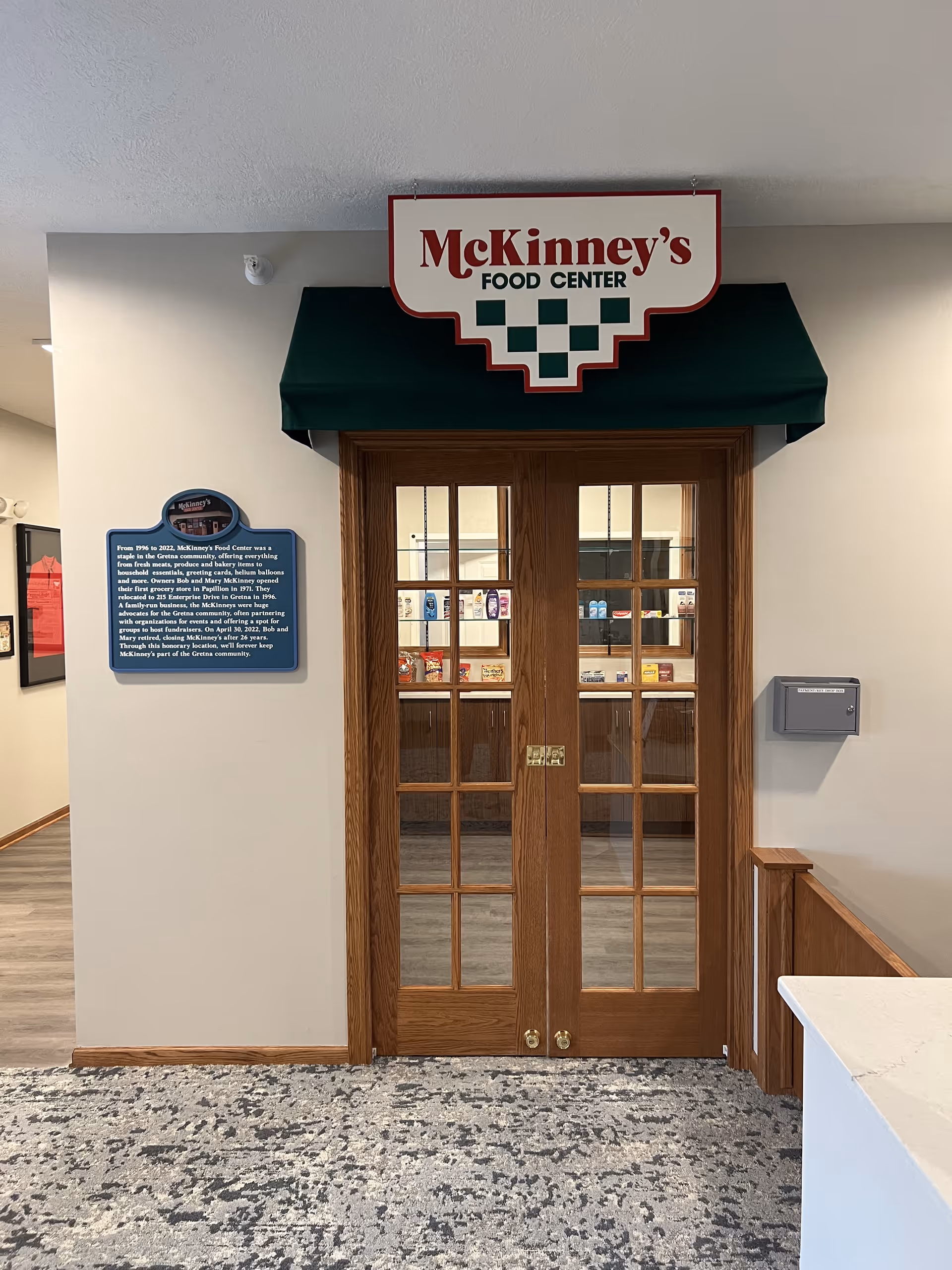 Double wooden doors with glass panes leading to McKinney's Food Center, with a green awning and a sign above the doors. To the left of the doors is a blue plaque with text, and to the right is a gray mailbox mounted on the wall. The floor is carpeted with a patterned design.