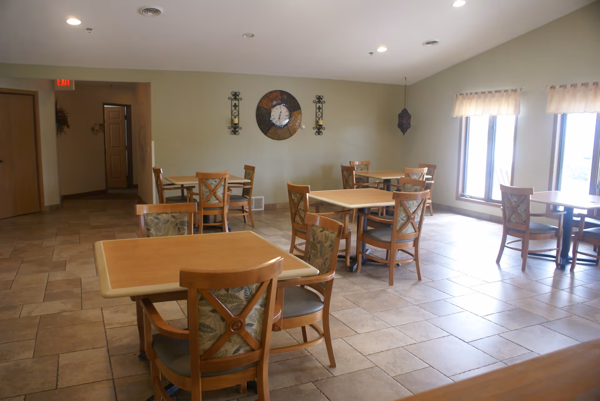 A dining room with several wooden tables and chairs arranged neatly on a tiled floor. The walls are light-colored with a decorative clock and candle holders mounted on one wall. Large windows with sheer curtains allow natural light to fill the room.