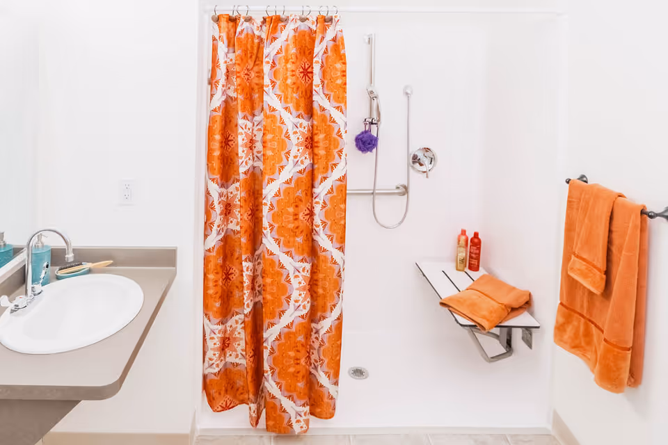 A clean bathroom with a white sink and countertop on the left, an orange patterned shower curtain in the center, and a walk-in shower with a fold-down bench, orange towels, and shampoo bottles on the right.