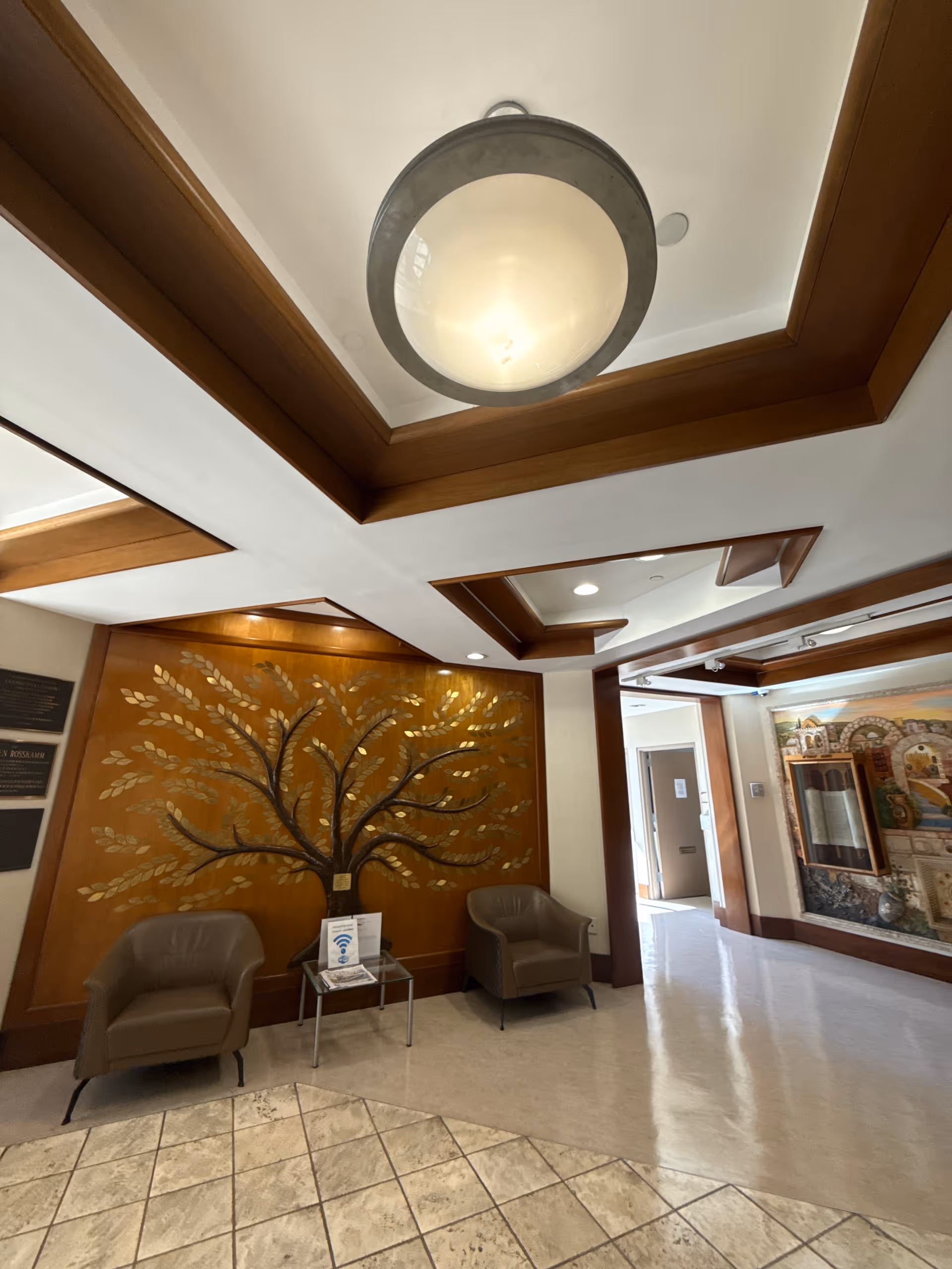 Interior view of a seating area in a nursing and rehabilitation center featuring two brown armchairs separated by a small glass table with a sign on it. Behind the chairs is a decorative wooden wall panel with a large tree design made of metal leaves. The ceiling has wooden beams and a large round light fixture. A hallway with artwork on the walls is visible to the right.
