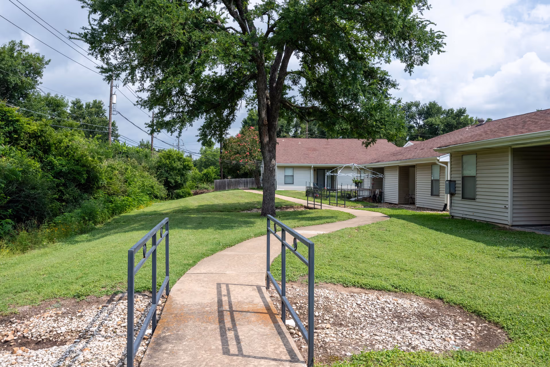 A paved walkway with metal railings leads through a grassy area with a large tree in the center. The path curves toward single-story buildings with beige siding and brown roofs, surrounded by green bushes and trees under a partly cloudy sky.
