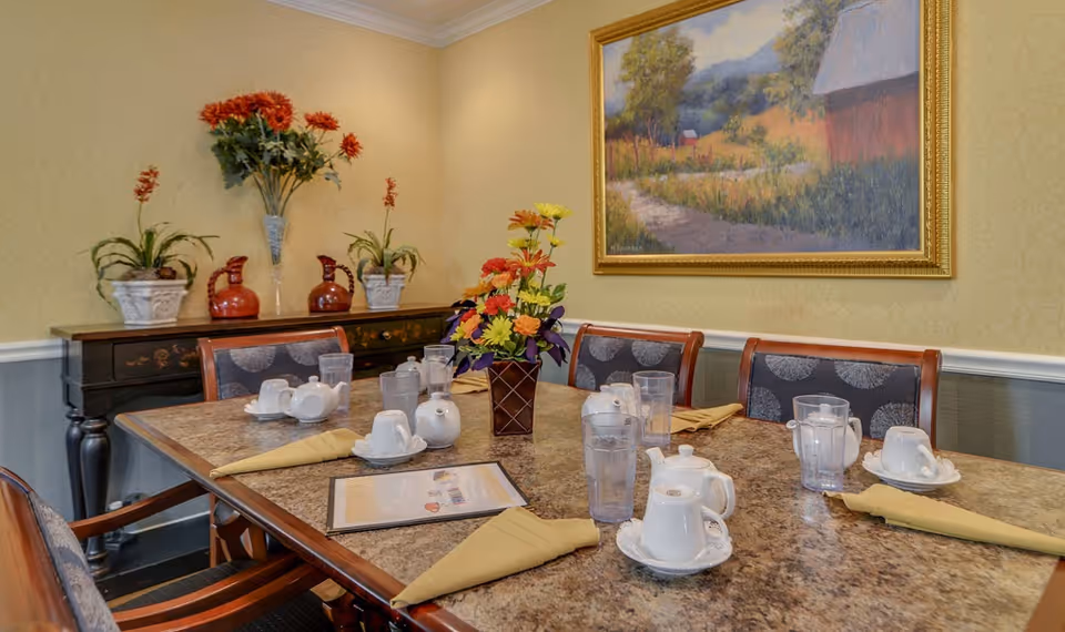 A dining room table set with white teapots, cups, glasses of water, and yellow folded napkins. A floral centerpiece is in the middle of the table. In the background, there is a sideboard with decorative plants and vases, and a framed painting of a rural landscape on the wall.