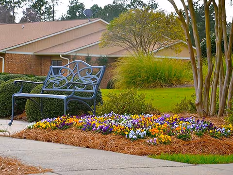 Decorative metal bench beside a colorful flower bed and landscaped lawn in front of a single-story building.