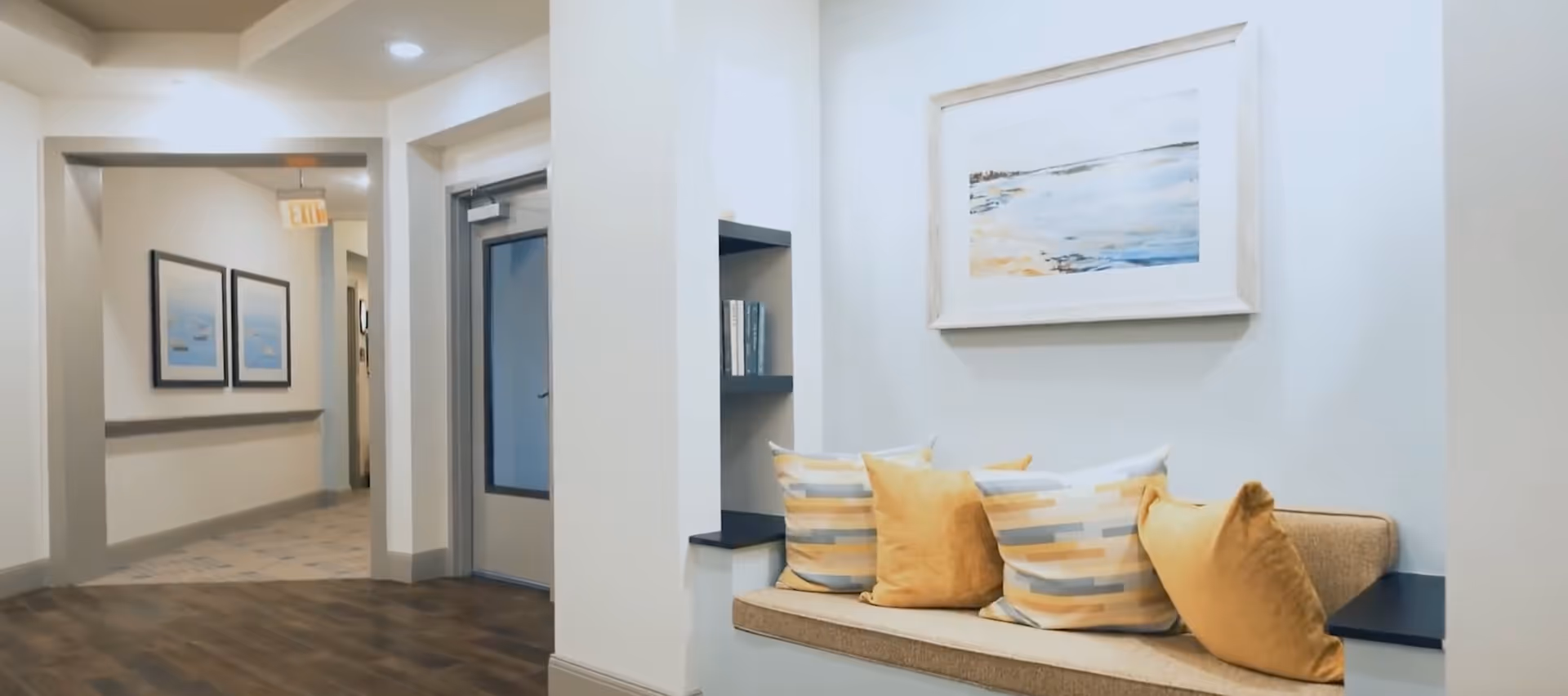 Bright interior hallway featuring a built-in bench with yellow and striped pillows, framed artwork, and a corridor leading past a door.