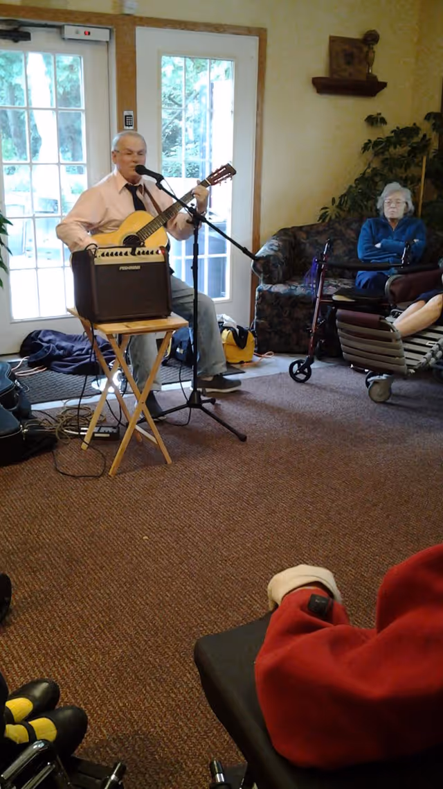 An elderly man is playing an acoustic guitar and singing into a microphone in a room with carpeted floor and glass doors. Several elderly people are seated around him, including a woman sitting on a couch with a walker nearby and another person in a wheelchair wearing a red jacket. There is an amplifier on a small wooden table in front of the man.
