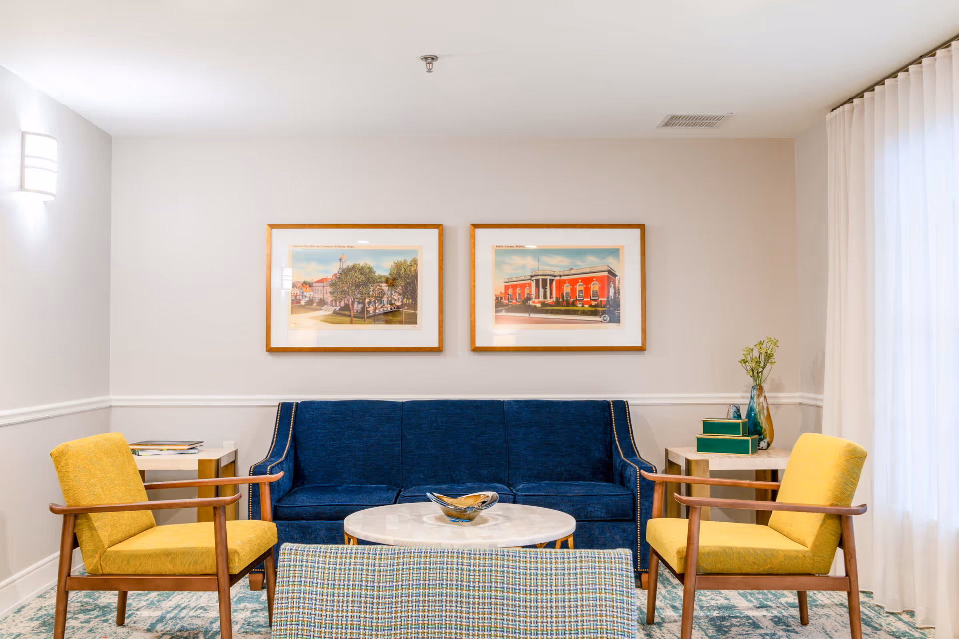 Bright sitting area with a blue sofa flanked by two yellow armchairs, a round marble coffee table, framed artwork on the wall, and side tables with decorative items.