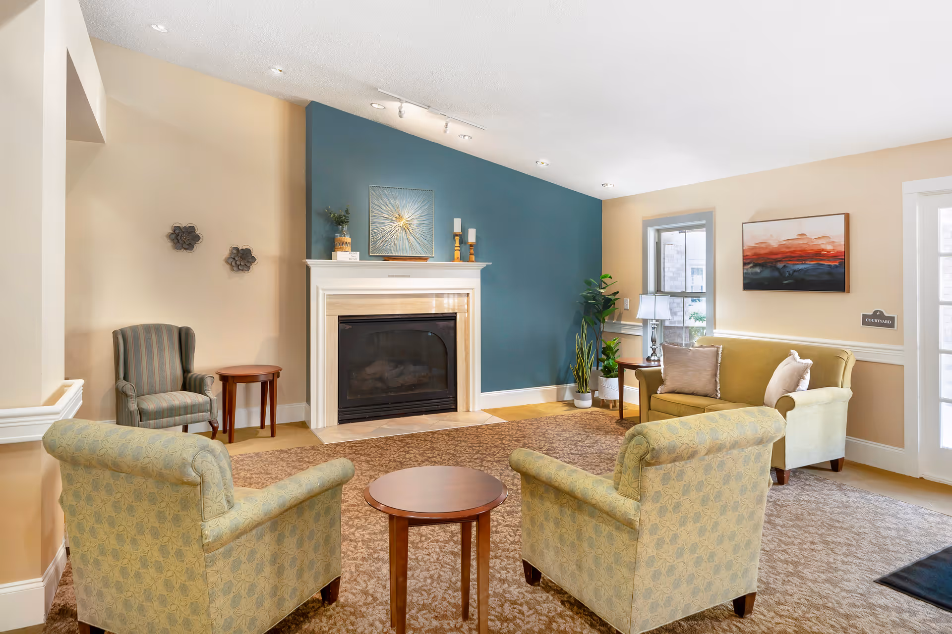A cozy living room area with a beige and blue color scheme featuring a fireplace with a white mantel, two patterned armchairs facing a small round wooden table, a green loveseat with pillows, a side table with a lamp, potted plants, and wall art. The room has carpeted flooring and a window letting in natural light.