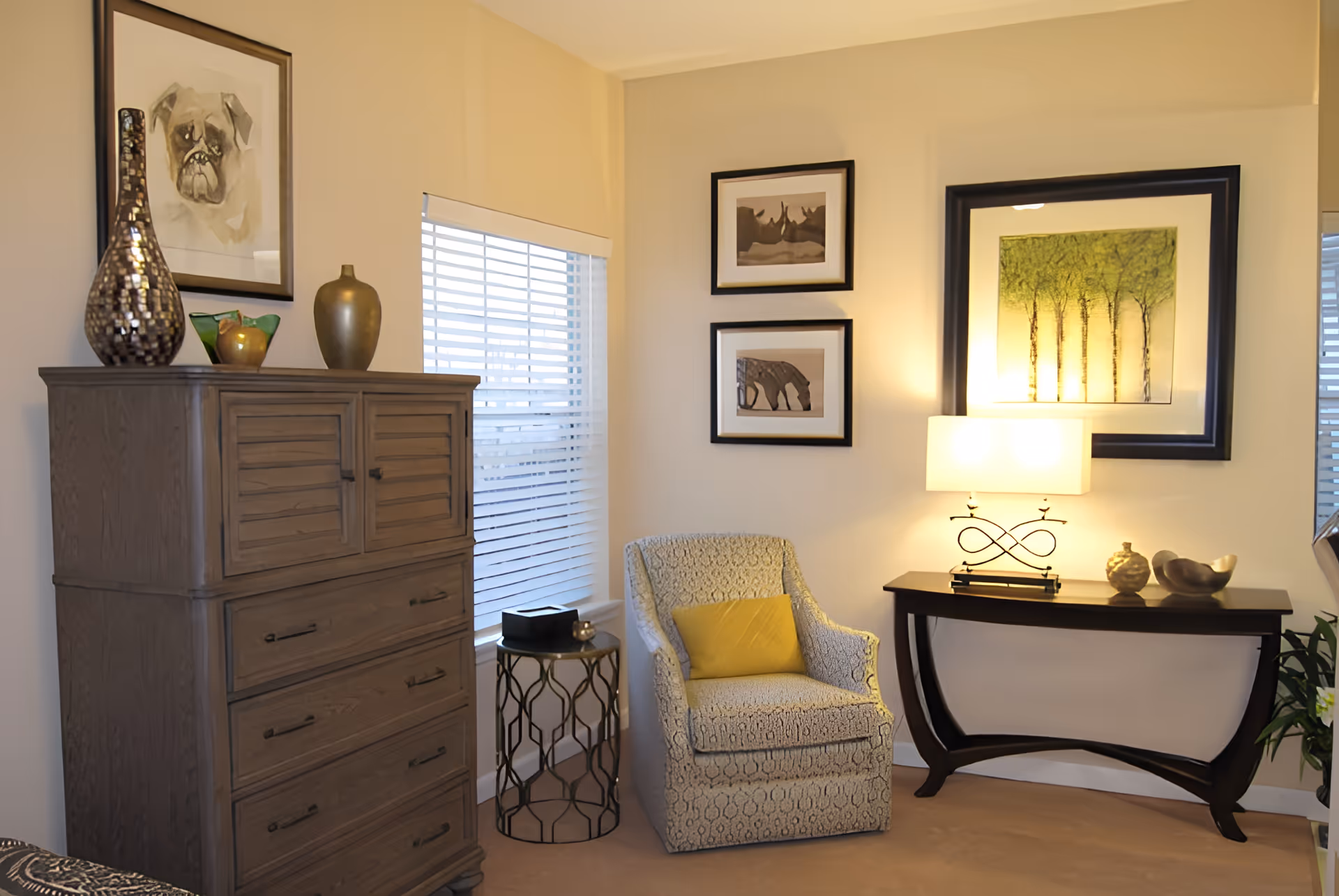 A cozy corner of a living room featuring a patterned armchair with a yellow cushion, a small round side table, a wooden chest of drawers with decorative vases on top, and a dark wooden console table with a lamp and decorative items. The walls are adorned with framed artwork including a painting of trees and animal sketches. A window with white blinds allows natural light into the room.