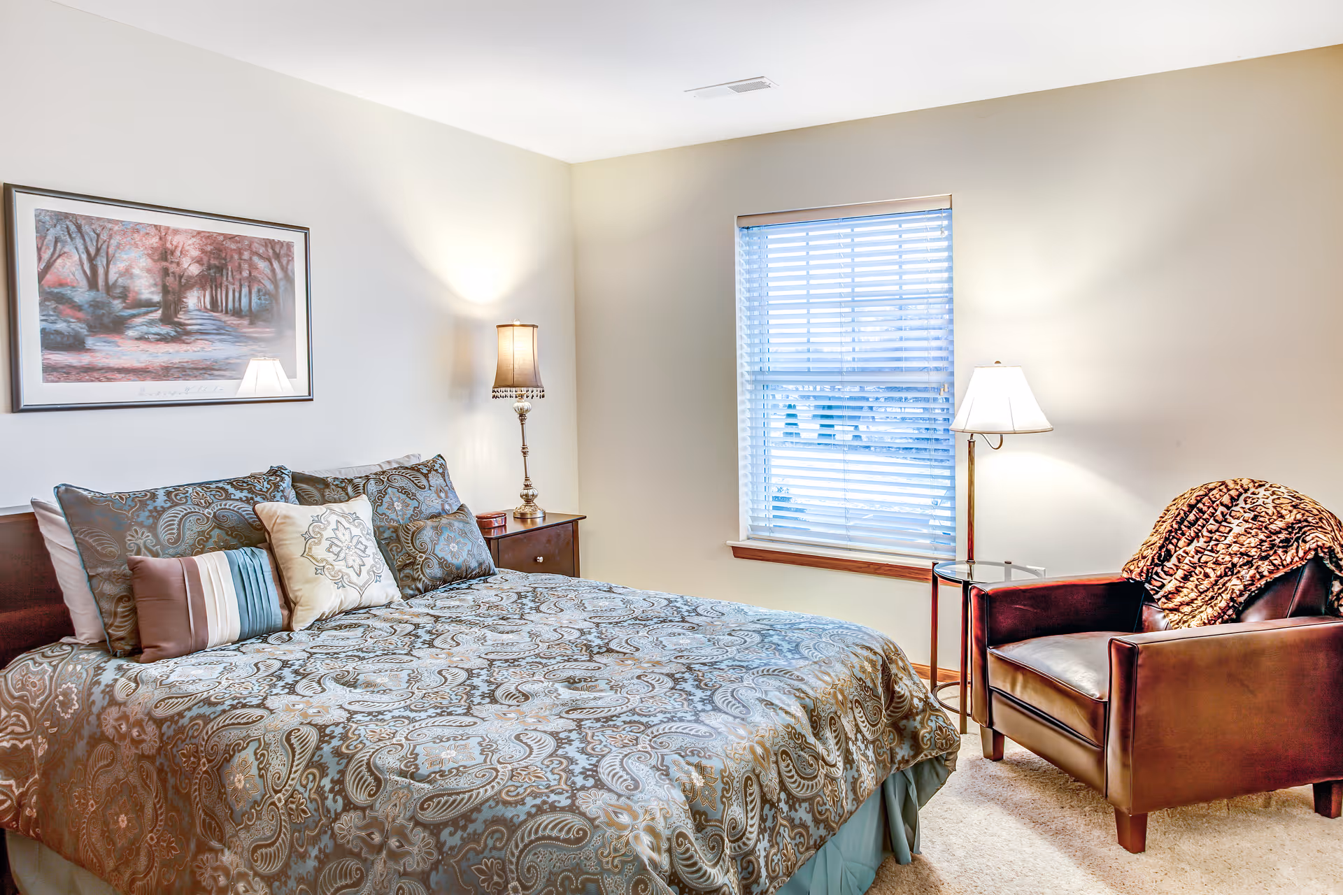 A cozy bedroom with a large bed covered in a patterned comforter and multiple pillows. There is a wooden nightstand with a lamp on the left side of the bed. A framed picture hangs on the wall above the bed. To the right, there is a window with blinds and a brown leather armchair with a patterned throw blanket draped over it. A floor lamp stands next to the armchair.