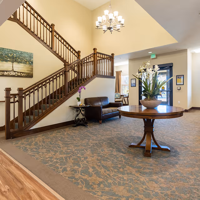 Lobby area with a wooden staircase, round table holding a planter of orchids, and leather seating near the entrance.