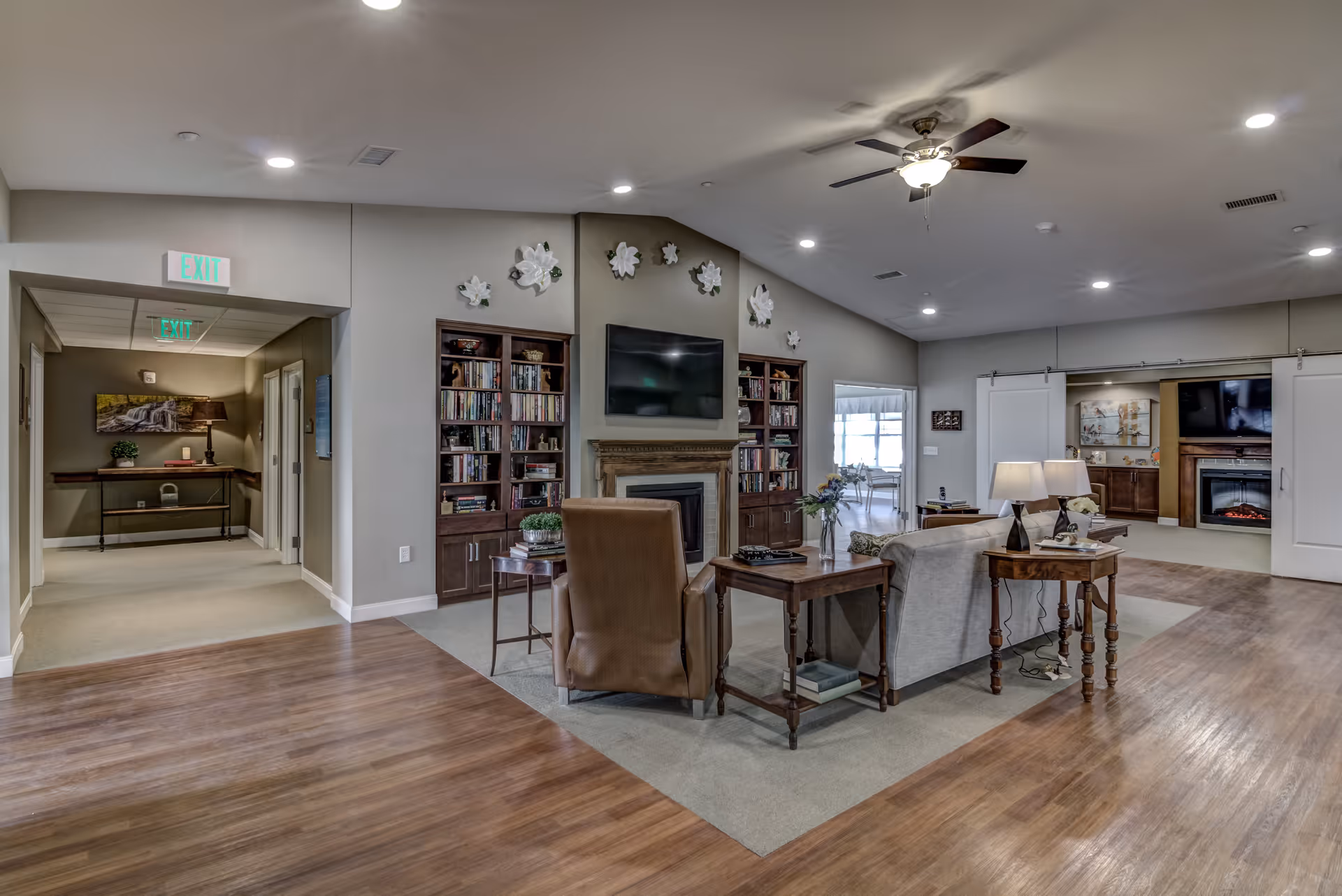 A spacious and well-lit living room area in a senior living facility with wooden flooring and carpeted seating area. The room features a ceiling fan with lights, a wall-mounted TV above a fireplace, built-in bookshelves filled with books, and comfortable seating including a sofa and armchair. Decorative white flowers adorn the wall above the fireplace. There are side tables with lamps and plants, and hallways leading to other rooms are visible.