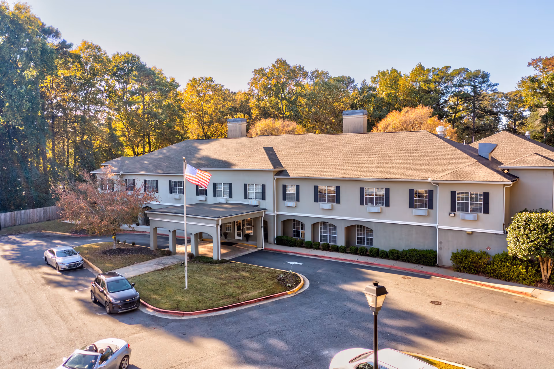 Exterior view of Greenwood Place Assisted Living and Memory Care building with a beige facade, multiple windows with black shutters, a covered entrance, an American flag on a flagpole, several parked cars, and surrounded by trees with autumn foliage.