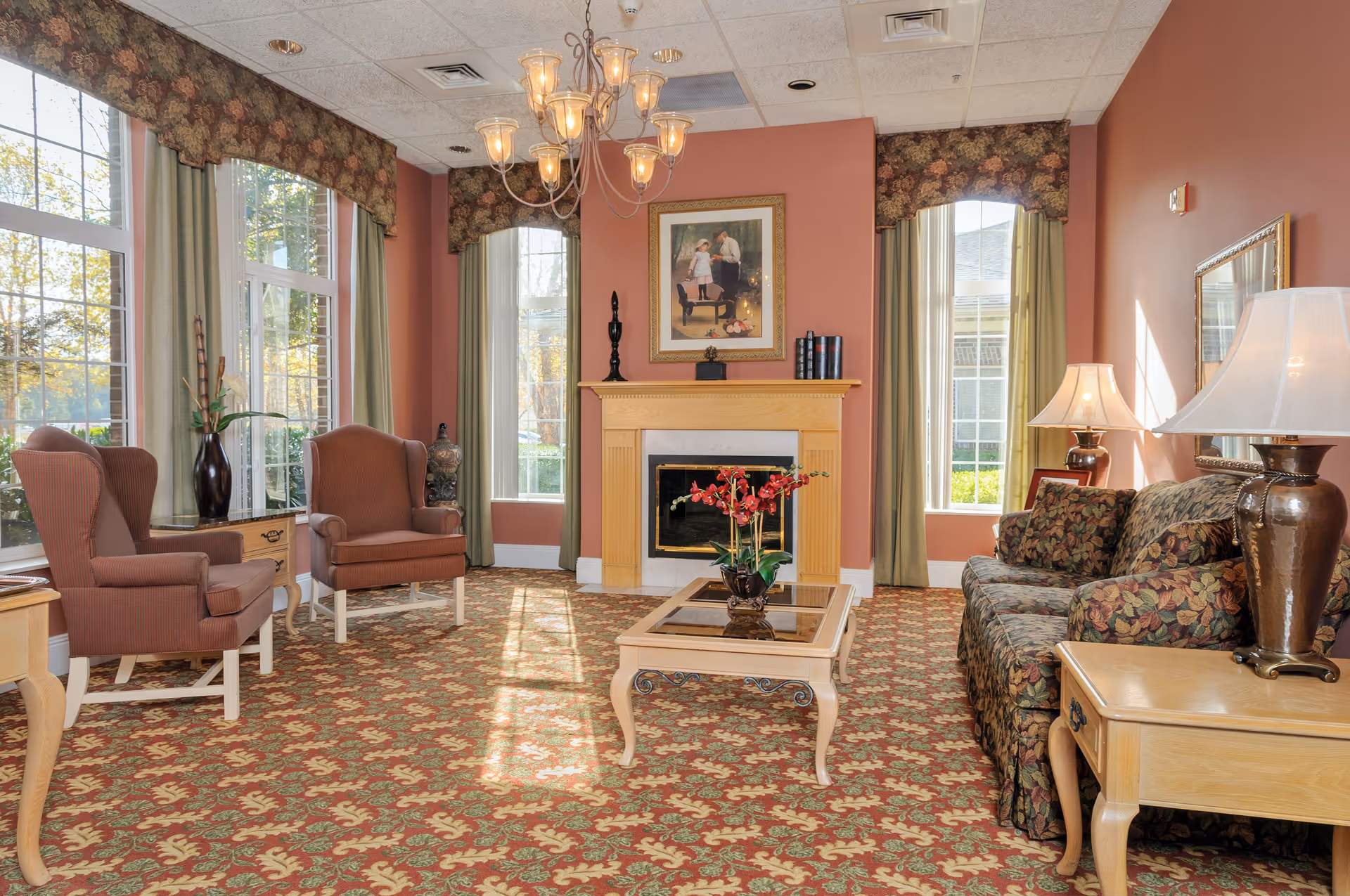 Bright lounge with patterned carpet, floral sofa, armchairs, coffee table, fireplace, and large windows.