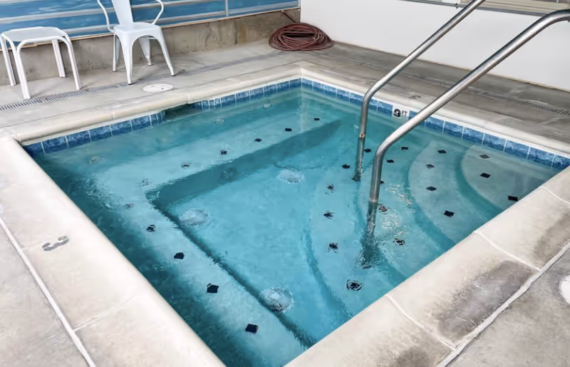 Indoor jetted hot tub with stainless steel handrail, tiled interior, and concrete deck with chairs nearby.