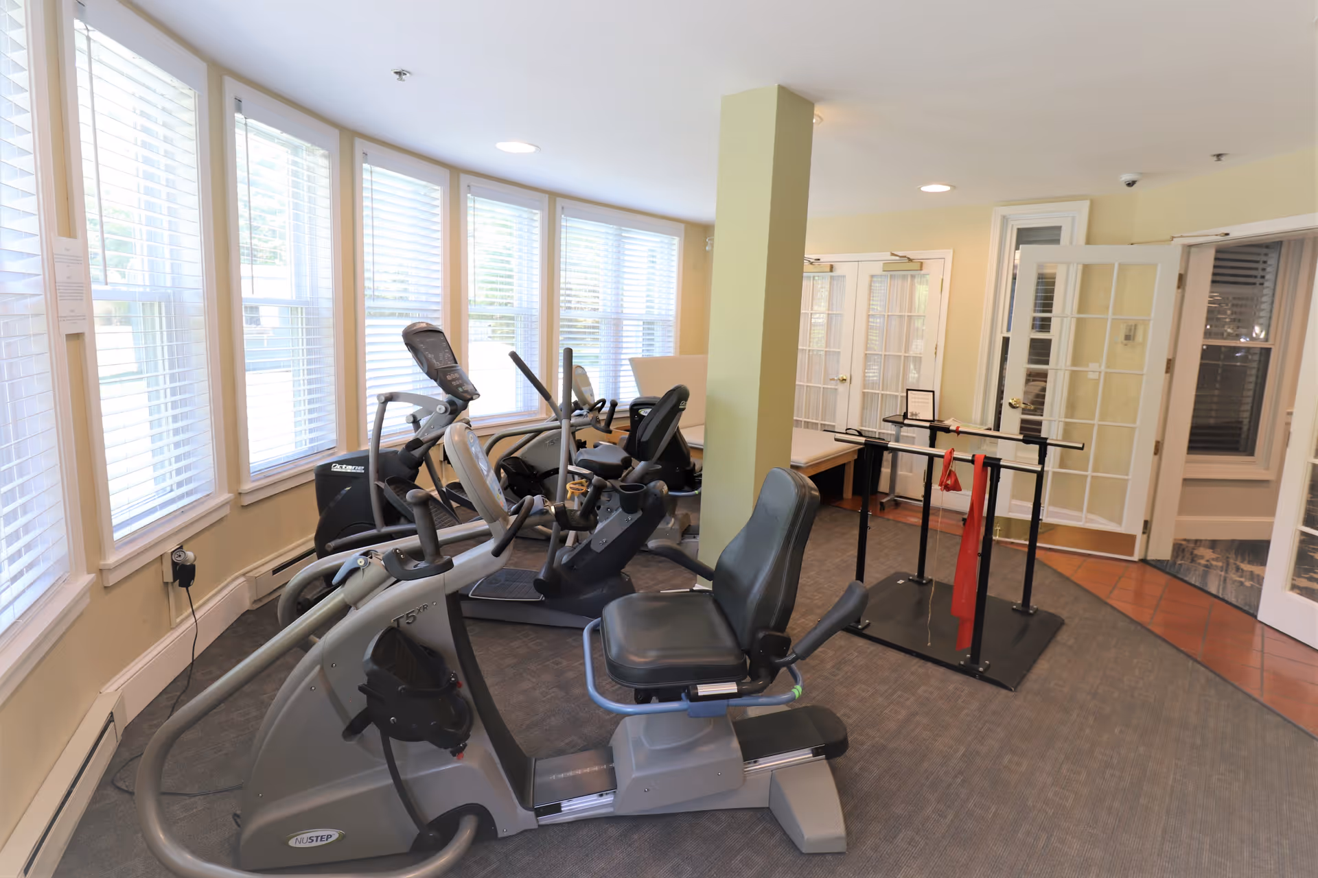 Small fitness room with recumbent exercise bikes, parallel bars, and a wall of windows.