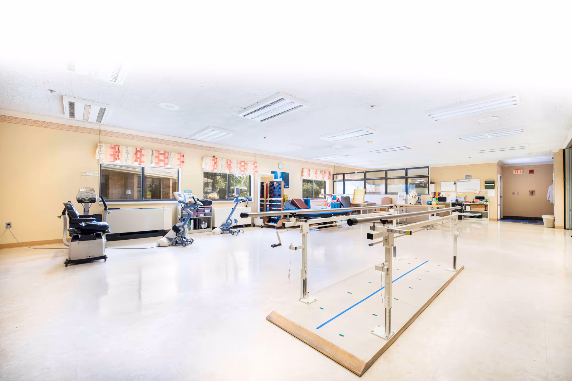 A spacious rehabilitation room with parallel bars for walking exercises in the center, exercise bikes along the left wall, and various physical therapy equipment and mats in the background. The room has large windows with patterned valances, beige walls, and bright overhead lighting.