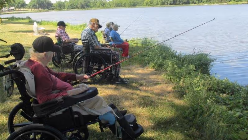 Four elderly individuals in wheelchairs fishing by the edge of a lake on a grassy area with trees in the background.