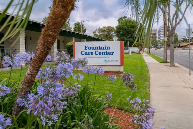 Outdoor view of the Fountain Care Center sign with purple flowers and a palm tree in the foreground, a sidewalk and street on the right, and buildings in the background under a cloudy sky.