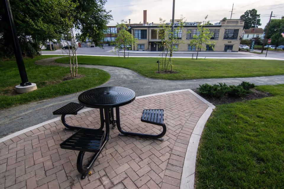 Outdoor seating area with a black metal round table and attached benches on a paved surface, surrounded by green grass, small trees, and walking paths. In the background, there is a two-story building with large windows and a street in front.