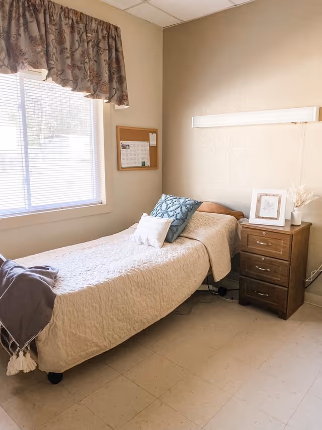 A small, simple bedroom with a single bed covered in a beige quilt and two pillows, one blue and one white. Next to the bed is a wooden nightstand with three drawers, a framed picture, and a small vase with dried flowers. A window with blinds and a floral valance lets in natural light. A cork bulletin board is mounted on the wall above the bed.