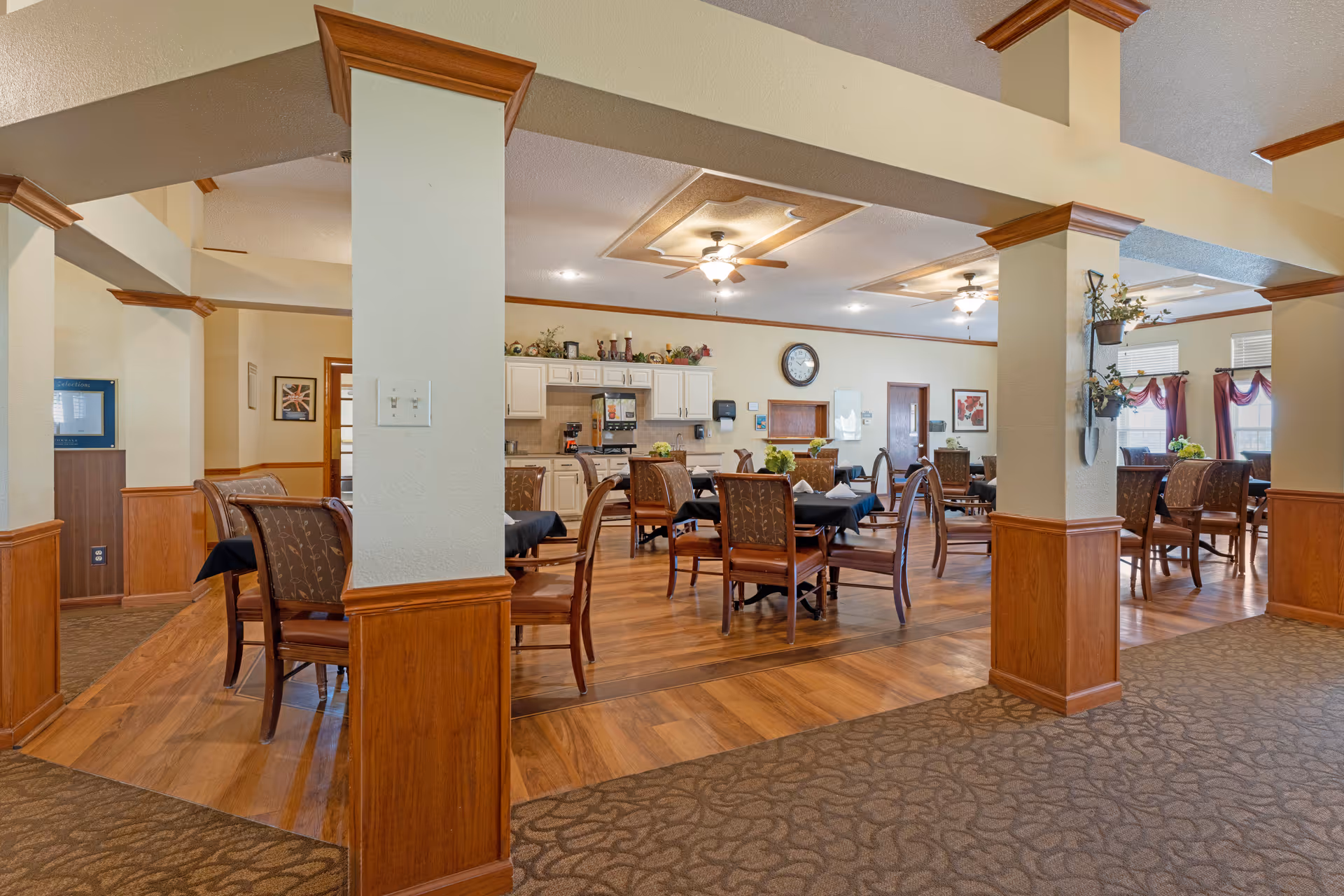 Dining room with multiple tables and chairs, wood columns and flooring and a kitchen area in the background.