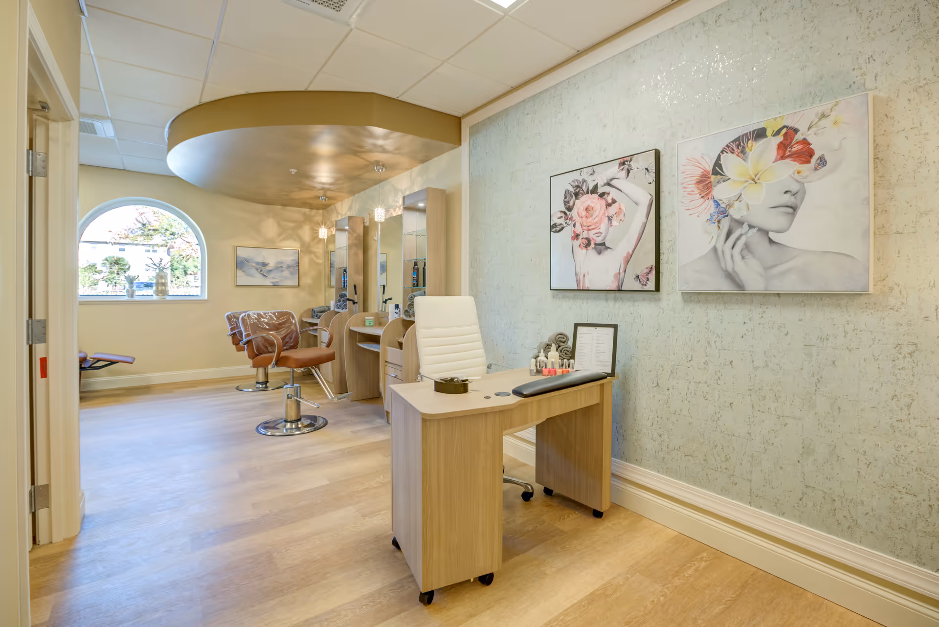 Interior view of a salon area in HarborChase of Daytona Beach featuring a manicure table with a white chair, nail polish and manicure tools on the table. In the background, there are two brown salon chairs in front of mirrors with shelves, under a curved ceiling section with pendant lights. The walls are decorated with artistic paintings of women with floral designs. A large arched window lets in natural light.