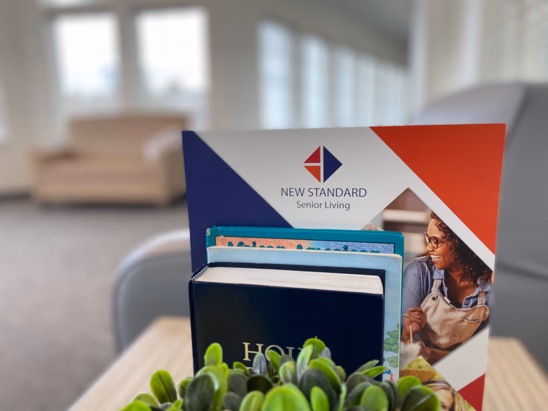 A close-up of a brochure holder on a table containing several books and brochures, with a New Standard Senior Living brochure prominently displayed. In the background, there is a blurred view of a beige couch and large windows letting in natural light.