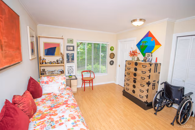 A bright and cozy room with light-colored walls and wooden flooring. There is a floral-patterned couch with red pillows on the left side, a small wooden shelf with decorative items, a red chair in front of a window with white blinds, a wooden chest of drawers with black handles, and a wheelchair positioned near the chest. Colorful artwork is displayed on the walls, and the room has a warm, inviting atmosphere.