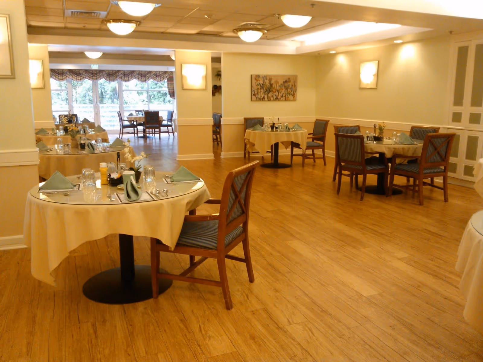 A well-lit dining room with several round tables covered with beige tablecloths. Each table is set with glasses, silverware, napkins, and small flower arrangements. Wooden chairs with blue cushioned seats surround the tables. The room has wooden flooring, soft yellow walls, and ceiling lights. Large windows with patterned valances allow natural light to enter the space.