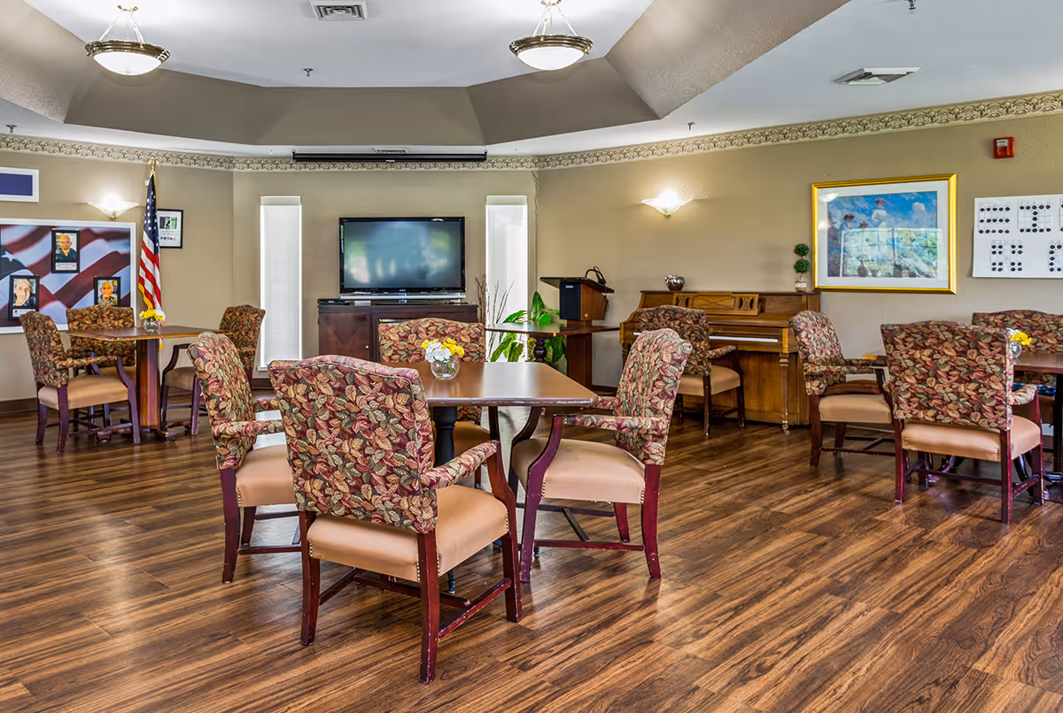 A common area in a senior living facility with several tables and chairs featuring floral upholstery. The room has wood flooring, a piano against one wall, a flat-screen TV, and framed pictures on the walls. There is an American flag in the corner and ceiling lights providing illumination.