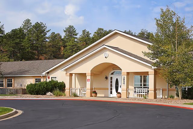 Single-story beige senior living facility entrance with a covered portico, double doors, potted plants and trees in the background.