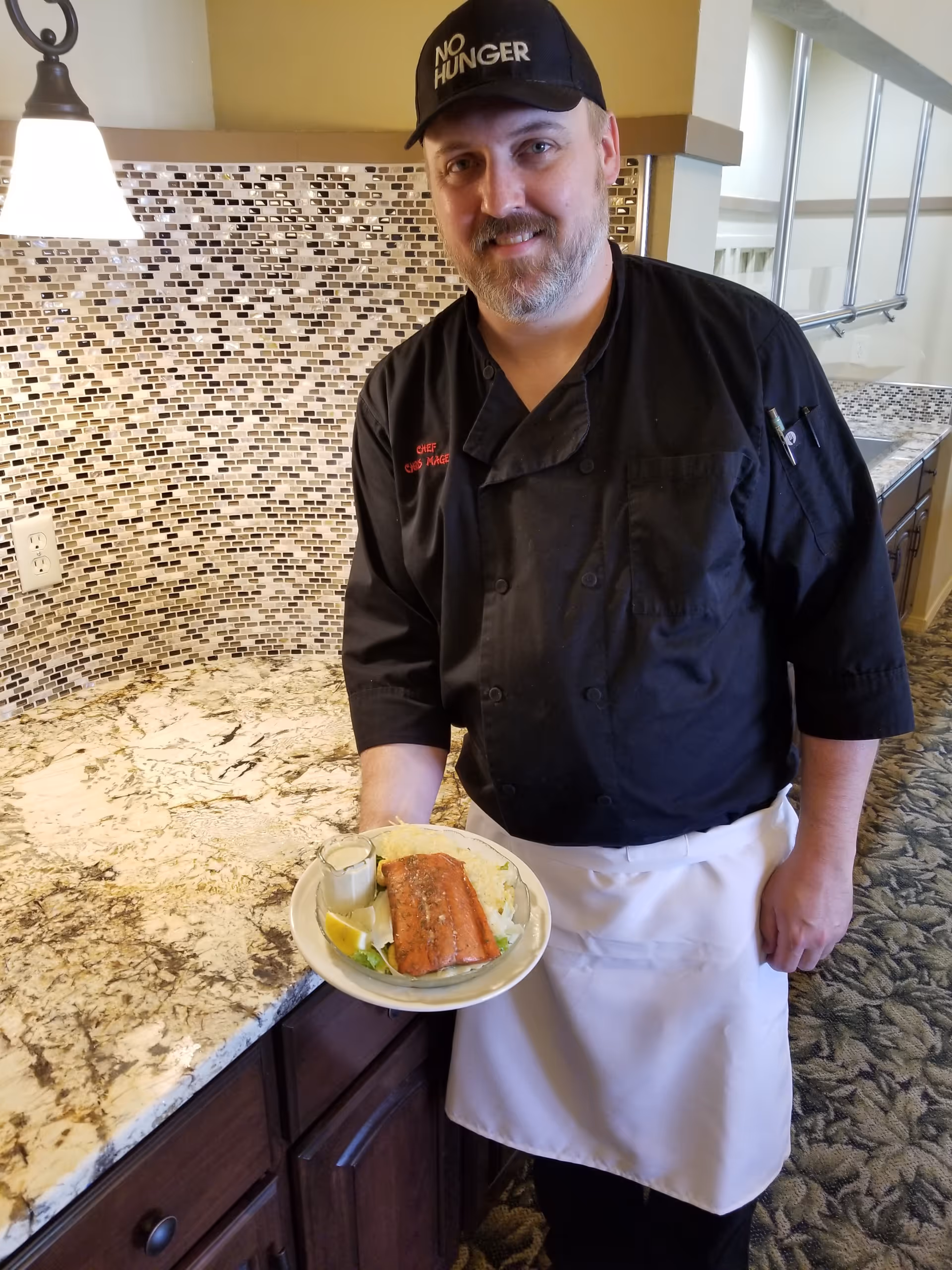 A chef wearing a black uniform and a black cap with the words 'NO HUNGER' stands in a kitchen area holding a plate with a cooked salmon fillet, mashed potatoes, and a small container of sauce. The background shows a tiled backsplash and granite countertop.