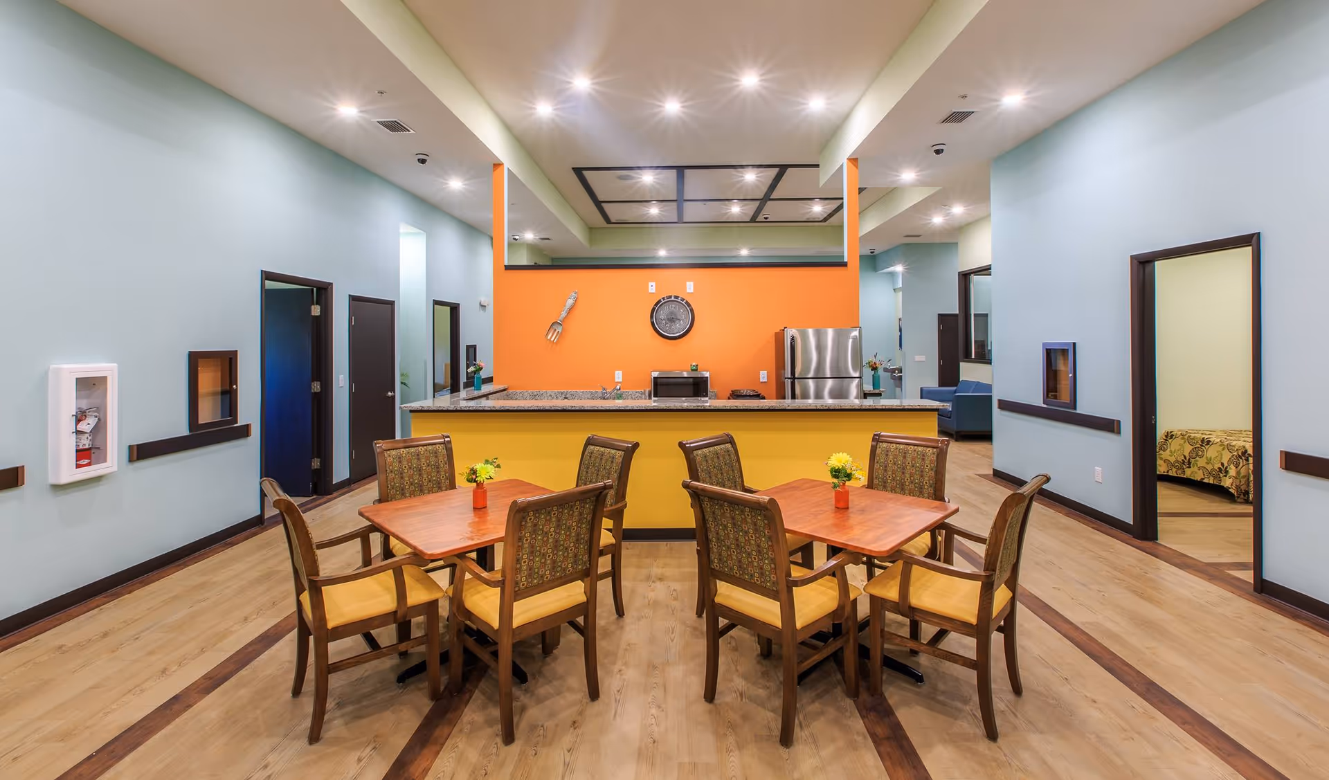 A bright and clean dining area in an assisted living facility featuring two wooden tables each surrounded by four chairs with patterned backs and yellow seats. The tables have small vases with yellow flowers. Behind the tables is a kitchen counter with a granite countertop, a microwave, a stove, and a stainless steel refrigerator. The wall behind the counter is painted orange and decorated with a clock and a large fork. The surrounding walls are light blue with several doorways leading to other rooms, including a bedroom with a floral bedspread visible on the right. The floor is wood with darker wood inlays, and the ceiling has recessed lighting.