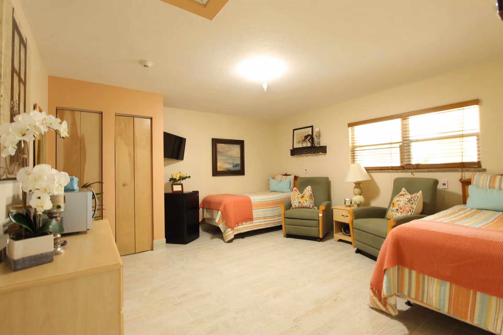 A bright and cozy senior living bedroom with two single beds, each with striped bedding and coral-colored throws. Between the beds are two green armchairs with floral pillows and a small wooden nightstand with a lamp. A window with wooden blinds lets in natural light. On the left side, there is a dresser with a microwave and decorative plants, and a wall-mounted TV above a small black cabinet. The walls are light-colored with framed artwork and a small shelf with decor.