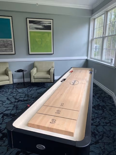 A recreational room with a shuffleboard table in the foreground, two green armchairs and a small round black table against a light blue wall with two framed abstract paintings. Large windows on the right side let in natural light.