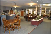Interior view of a senior living facility common area with multiple wooden tables and chairs, a reception desk in the background, potted plants in a raised planter, and columns wrapped with decorative material.