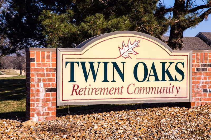 A large outdoor sign for Twin Oaks Retirement Community made of brick and a cream-colored panel with green and red lettering, set against a backdrop of trees and grass.