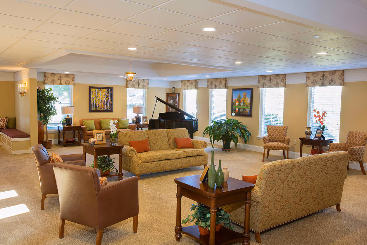 Communal living room with sofas, armchairs, decorative plants and a grand piano under recessed lighting.