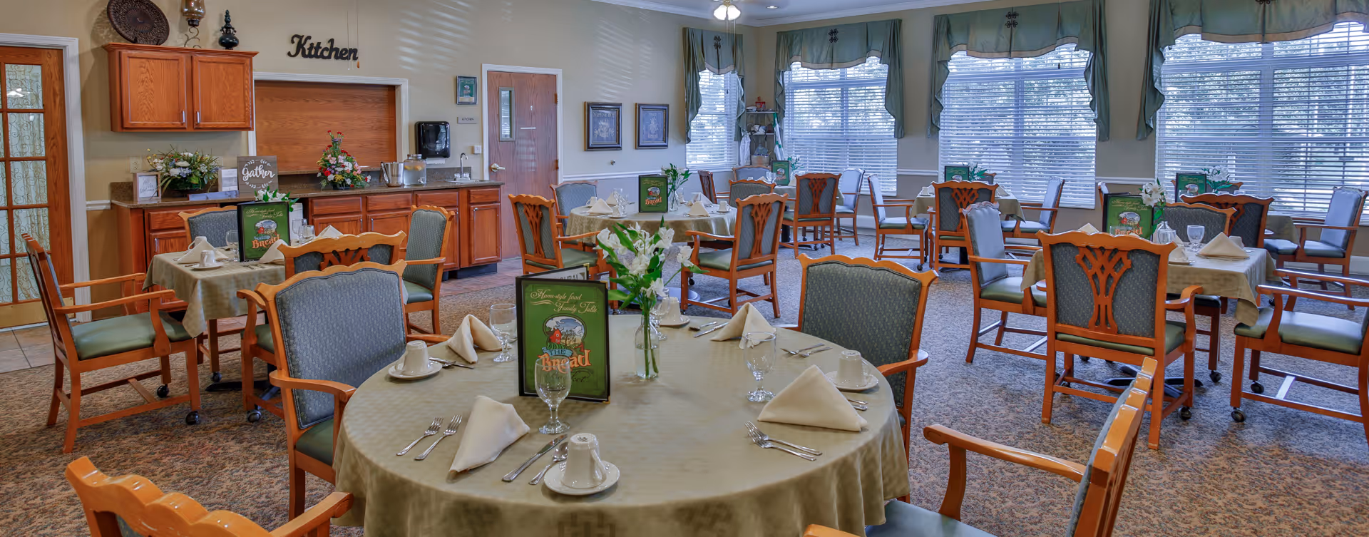 A dining room with multiple round and square tables set with beige tablecloths, folded napkins, cups, glasses, and silverware. The room has wooden chairs with green cushions, large windows with green valances, and a sideboard with flowers and a sign that says 'Kitchen' above it.